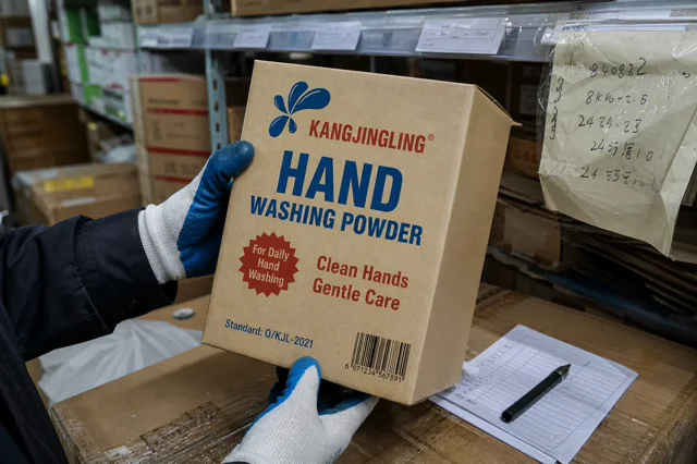 Qiaoshou hand washing powder carton held by a gloved worker in a wholesale store back room with shelf notes and a sample check sheet